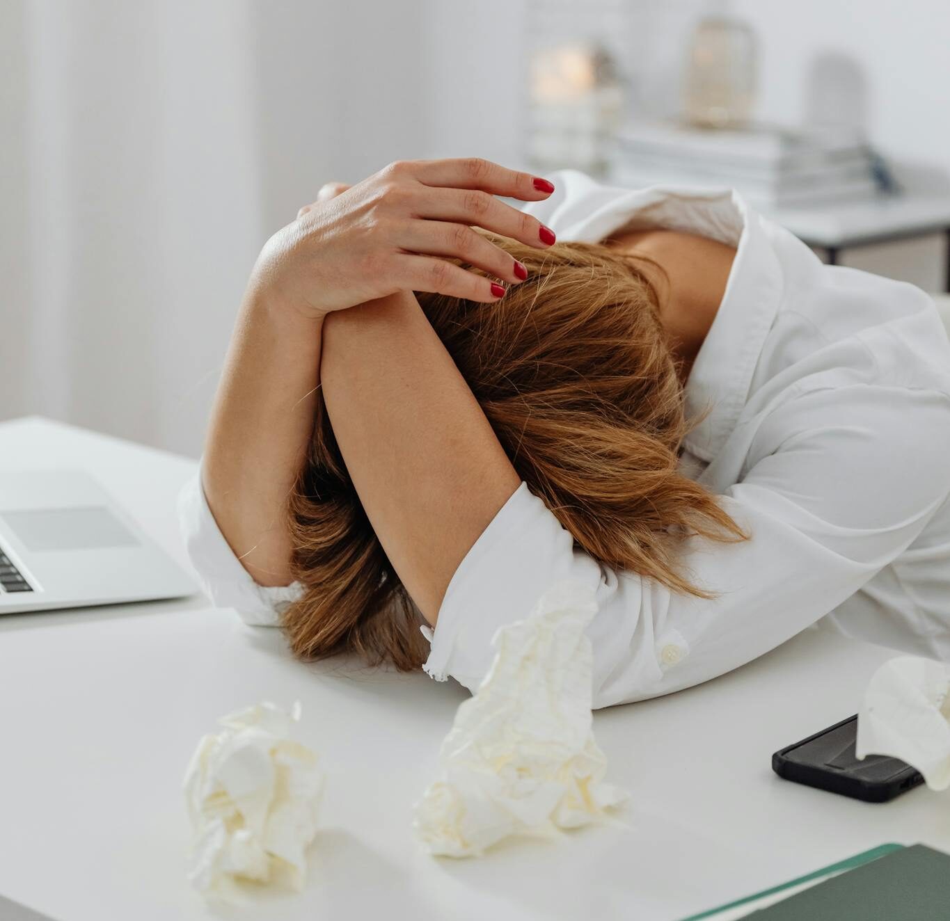 A woman in distress sits at a desk with head in hands, surrounded by tissue papers.